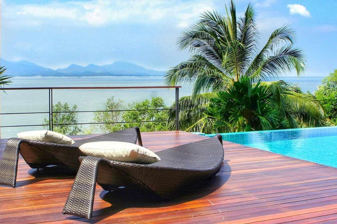 Modern timber deck with rattan lounge chairs beside an infinity pool overlooking the ocean and tropical palm trees.