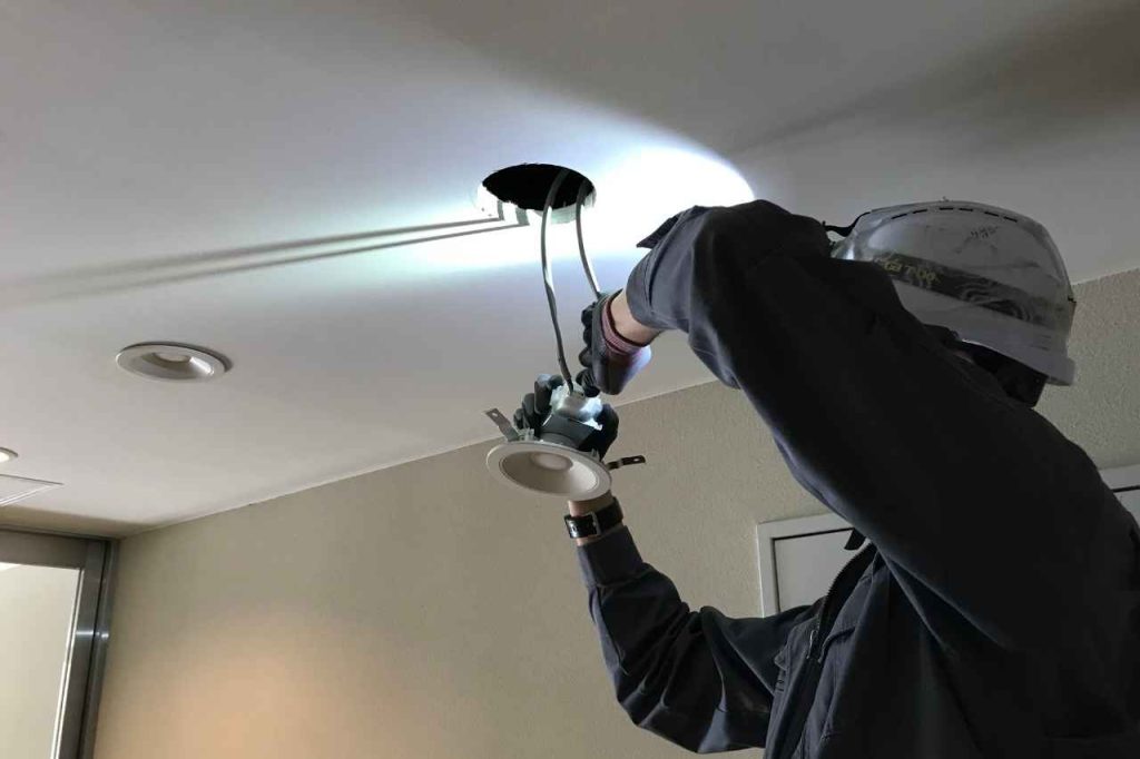 Licensed electrician from CAESA installing a recessed ceiling light fixture, wearing safety helmet and gloves, performing electrical wiring work indoors under bright LED light.
