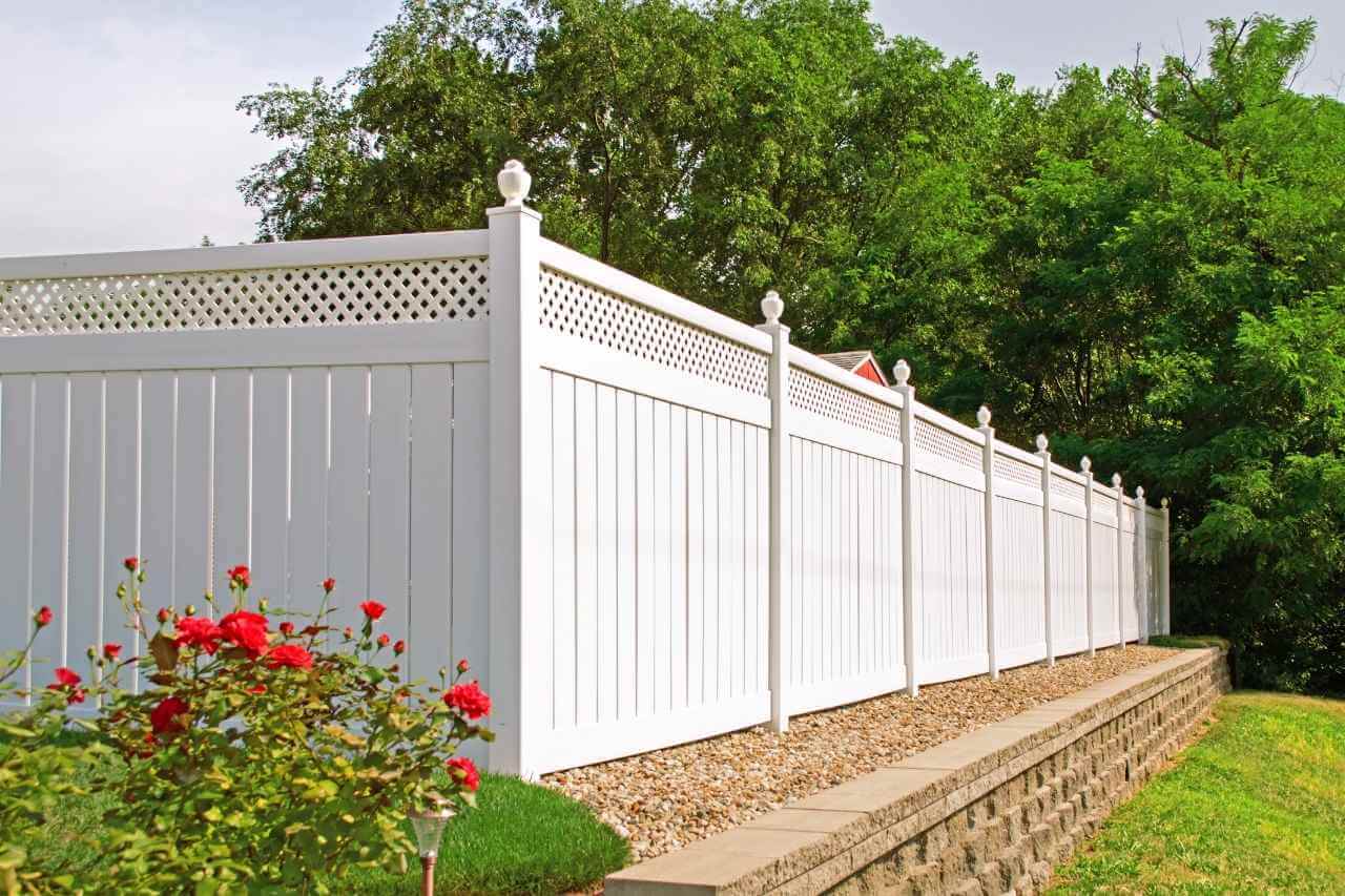 White vinyl privacy fence with decorative lattice top installed along a landscaped garden with red roses and green trees.