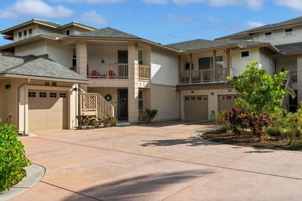 Modern residential house with beige exterior and large driveway made of light-colored decorative concrete, surrounded by greenery and blue sky.