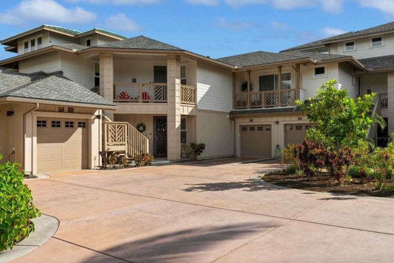 Modern residential house with beige exterior and large driveway made of light-colored decorative concrete, surrounded by greenery and blue sky.