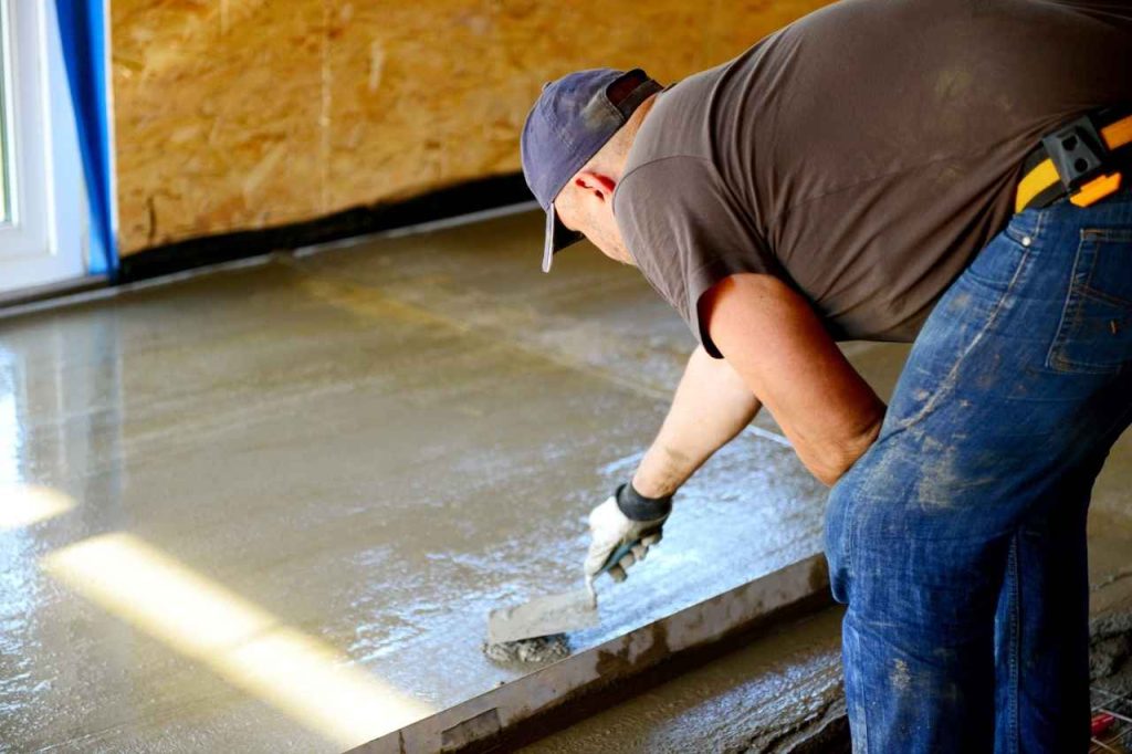 Worker smoothing freshly poured concrete on an indoor floor using a hand trowel, wearing gloves, jeans, and a cap during the finishing process.