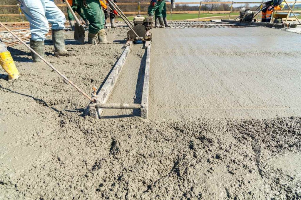 Construction workers leveling freshly poured concrete using a power screed machine on a reinforced slab, wearing safety boots and workwear at a job site.