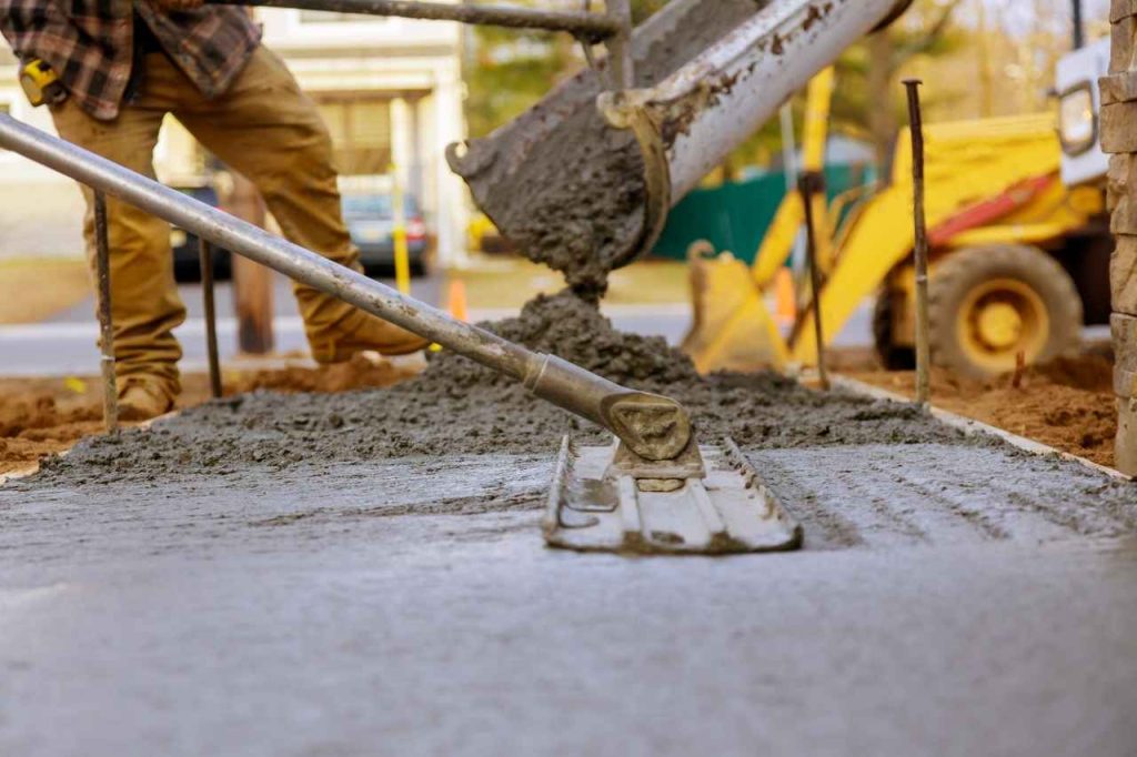 Worker spreading and smoothing freshly poured concrete on a driveway using a hand float, with concrete being poured from a chute in the background.