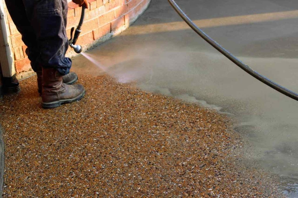 Worker cleaning and revealing exposed aggregate concrete surface using a water hose near a brick wall during the finishing process.
