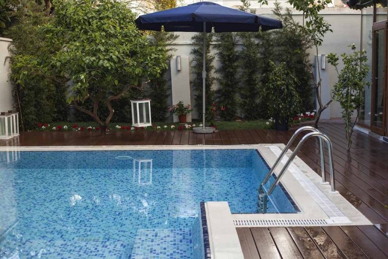 Modern backyard plunge pool with mosaic tiles, wooden deck, and garden landscaping featuring trees and outdoor umbrella.